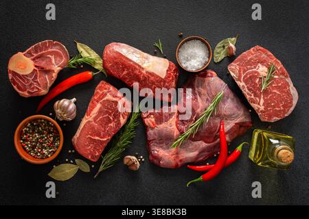Various cuts of beef steak are shown with herbs, spices, and oil on a dark surface. Perfect for those looking to prepare or grill different beef dishes at home Stock Photo