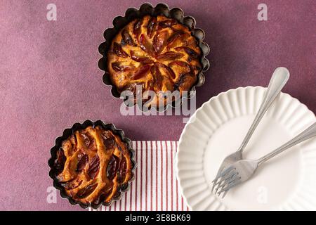 A plum pie is placed on a table with thinly sliced plums visible on top. Bowls of fresh plums are nearby with empty plates waiting for dessert Stock Photo