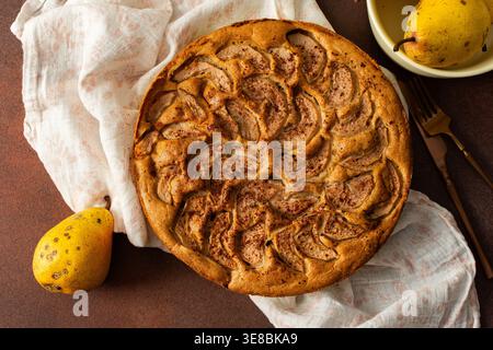 A fresh pear pie is placed on a cloth on a table. Nearby, a bowl holds ripe pears and a small vase has flowers. The setting looks warm and inviting for sharing a treat Stock Photo