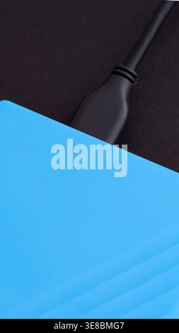 A close-up of a blue external hard drive with a black USB cable connected to it. Black background. Stock Photo