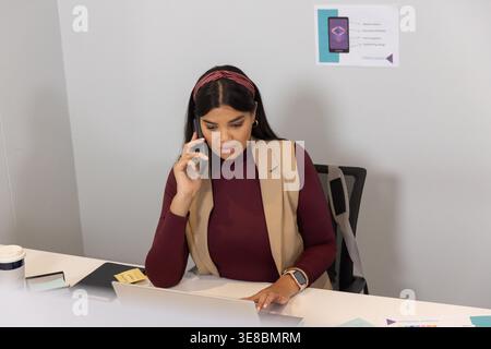 Mid-adult female in burgundy top and beige blazer holding smartphone to ear, using laptop at desk Stock Photo