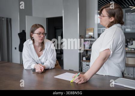 Businesswomen discussing and exchanging papers at desk in bright company office during work meeting. Discussing, exchanging, desk, office Stock Photo