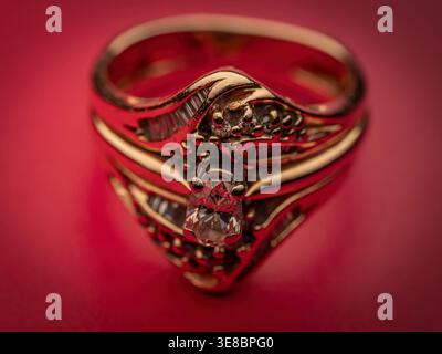 Macro photograph of a gold diamond ring with a large center round cut diamond and smaller baguette side stones on a dark red textured surface Stock Photo