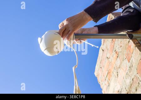 A man's hands install a white plastic CCTV camera on a metal bracket on a brick wall under a blue sky. Home security, outdoor technology, DIY installa Stock Photo