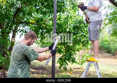Two workers collaborating to install a metal fence pillar in a sunny garden. Teamwork, manual labor, construction site, garden project. Stock Photo