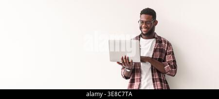 Young Man Using Laptop While Standing in Front of a White Wall in a Casual Setting Stock Photo