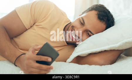 Man Lying on Bed Using Smartphone and Smiling While Looking at Screen During Daytime Stock Photo