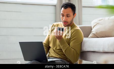 Man Using Voice Command on Smartphone While Working on Laptop in Living Room Space Stock Photo