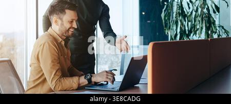 Two Coworkers Work Together in an Office Setting With a Laptop and a Bright Window Stock Photo