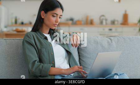 Serious concentrated Indian woman working studying online typing laptop at home looking away thinking pondering Arabian female girl author writing PC Stock Photo