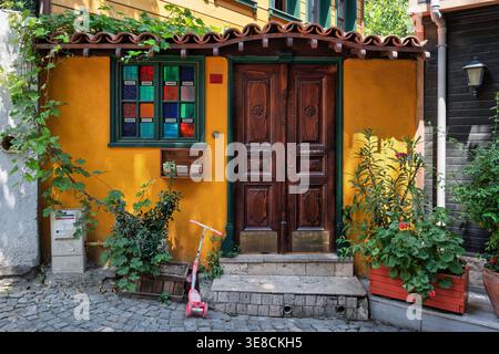 Vibrant yellow house facade in Istanbul, Turkey. Features a carved wooden door, colorful stained glass window, terracotta roof, lush plants, and a pink scooter on cobblestones Alley Stock Photo
