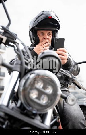 Motorcycle rider reacting to shocking news on phone, showing intense surprise expression. Distracted driver concept Stock Photo