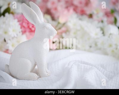 One white bunny figurine Easter Spring statue on soft fabric with blurry pink and white flower floral blossoms background Stock Photo