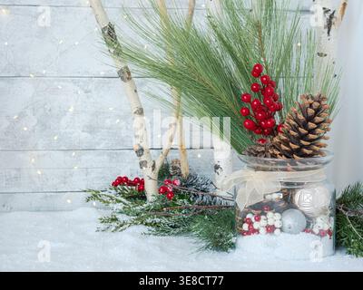 Christmas glass jar with ornaments and pinecones on snow ice, birch tree branches and festive holiday twinkle fairy lights, greenery and red berries Stock Photo