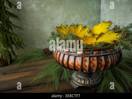 Fall gold leaves and spruce branches in a brown festive carved wood pedestal bowl on autumn rustic plank table with long pine needles on muted green Stock Photo