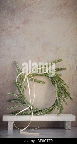 Background for personalized Christmas card. Vertical boho chic Norfolk pine wreath, white ribbon bow on weathered wood white stand rustic background Stock Photo