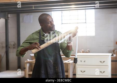 African American man standing in woodshop wearing dark work apron measuring plank with tape measure Stock Photo