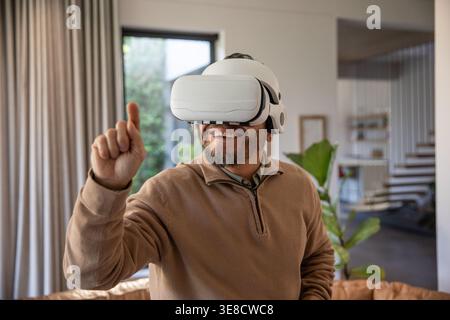 Mature Asian man standing in living room wearing white VR headset, pointing at virtual interface Stock Photo