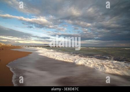 Waves crash on the beach near the pier during sunset Stock Photo