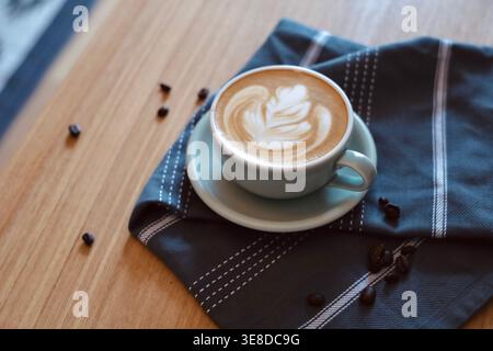 Latte art leaf design in a sage green ceramic cup on a dark plaid cloth surrounded by scattered coffee beans. A warm cozy cafe aesthetic capturing artisan barista craft and lifestyle comfort. Stock Photo