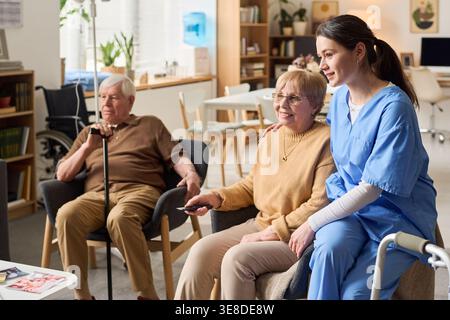 Senior Caucasian woman sitting with female caregiver, using remote control while senior man resting nearby, representing assisted living support. Ideal for elder care marketing, healthcare services Stock Photo