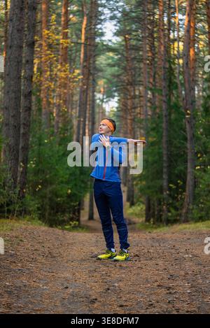 Young athletic man stretching his shoulder muscles before a workout, standing on a forest trail surrounded by tall pine trees, focusing on fitness and wellbeing in nature Stock Photo