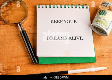 A white notepad with KEEP YOUR DREAMS ALIVE sits beside a magnifying glass and rolled US 100 dollar bills on a wooden surface, suggesting planning. Stock Photo