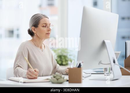 Mature, woman and writing with computer in office for journalism, online report or schedule. Female person, research or journalist taking notes in Stock Photo