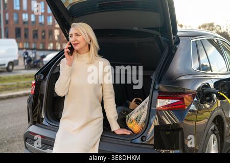 A professional woman speaking on her mobile phone while standing next to the open trunk of an electric vehicle in an urban environment. Stock Photo