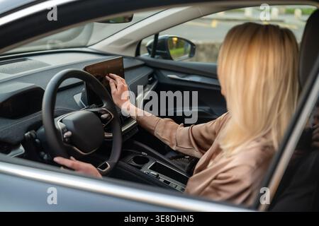 Female driver engaging with advanced touchscreen infotainment system inside a modern car. Interior features sleek design elements and ambient lighting enhancing the driving experience. Stock Photo