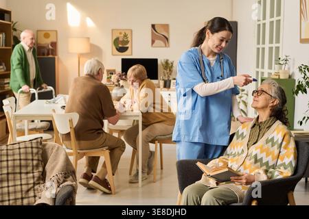 Senior woman receiving temperature check from female nurse while reading in care home, with elderly residents socializing behind. Ideal for eldercare, assisted living, healthcare support marketing Stock Photo