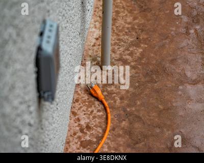 Shot shows a bright orange heavy duty extension cord lying unplugged on a wet, textured concrete Stock Photo