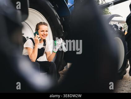 Woman farmer sitting on wheel of tractor and using her mobile phone. Stock Photo