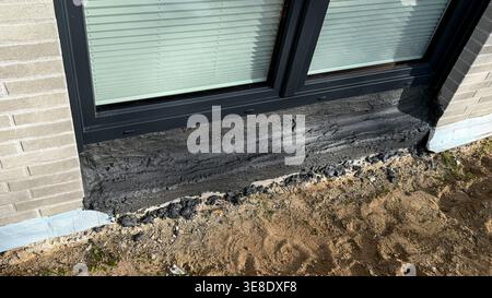 Close-up view of freshly applied black sealant around the base of a modern window on a residential building with sandy ground and brick wall visible Stock Photo