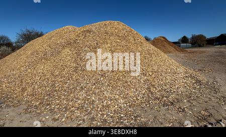 Large mound of wood chips in a sunny outdoor setting with clear blue sky and additional smaller piles visible in the background Stock Photo