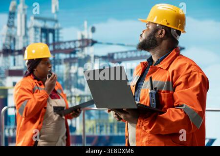 Team of offshore rig technicians monitoring operations using laptop and tablet on oil platform deck. Teamworking drilling rig coworkers reviewing energy operations using devices Stock Photo
