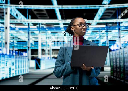 Data center female technician uses laptop program to conduct security audits and protect against cyber threats. African american woman uses notebook to implement cybersecurity measures in server farm Stock Photo