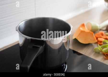 Empty cooking pot placed on a stovetop in a modern kitchen with fresh vegetables prepared on the counter. Food preparation process and home cooking co Stock Photo