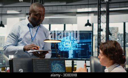 Manager talking with intern in office, using specialized hardware to accelerate complex AI calculations. Team leader teaching trainee how to manage machine learning training datasets, camera B Stock Photo