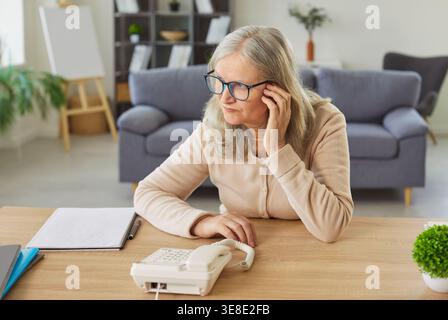 Worried sad senior woman sitting at the desk at home waiting for important phone call from someone. Stock Photo
