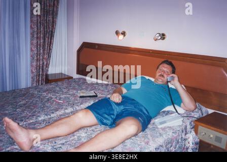 Man lying on hotel bed talking on landline phone 1990s Stock Photo