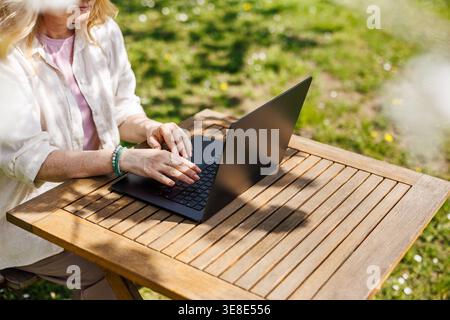 Freelancer working on laptop in spring garden. Woman using computer at outdoor desk in backyard. Remote work from home in sunny day Stock Photo