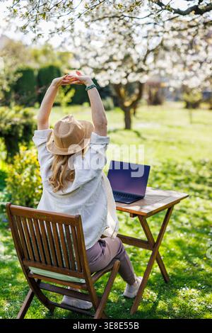 Woman stretching hands while working with laptop outdoors. Stress relief. Exercising body during remote work break in spring garden Stock Photo
