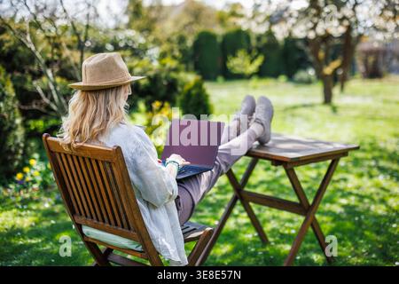 Woman working on laptop while sitting on chair with feet up on table in spring garden, enjoying freedom of remote job. Freelance work Stock Photo