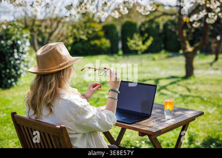 Woman putting on eyeglasses while working on laptop in spring garden. Female freelancer using computer outdoors during sunny day Stock Photo