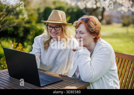 Multi-generation family using technology. Adult daughter teaching senior mother how to make video call on laptop while sitting at table outdoors Stock Photo