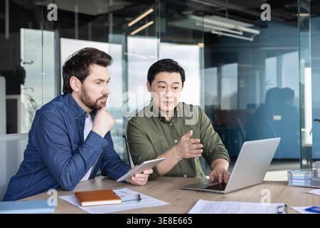 Two diverse business professionals collaborating during a discussion, reviewing information on a laptop and tablet, working together on a project in a bright contemporary office Stock Photo