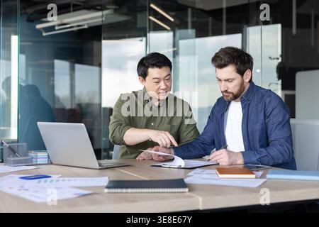 Two diverse businessmen are analyzing documents and discussing details around a desk in a modern office, working together on a strategic partnership or agreement Stock Photo