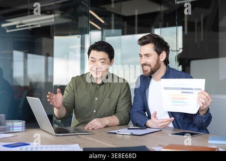 Two diverse businessmen are collaborating in an office, discussing sales projections and marketing data presented on a laptop and a printed graph, fostering teamwork and professional cooperation Stock Photo