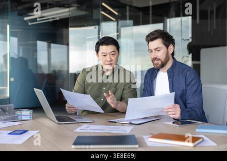 Two diverse business partners discussing a project, reviewing documents and data together at an office desk, highlighting teamwork and professional collaboration Stock Photo
